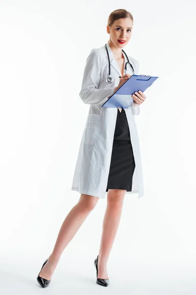 Infirmière heureuse et sexy en manteau blanc écriture prescription tout en tenant presse-papiers sur blanc — Photo de stock