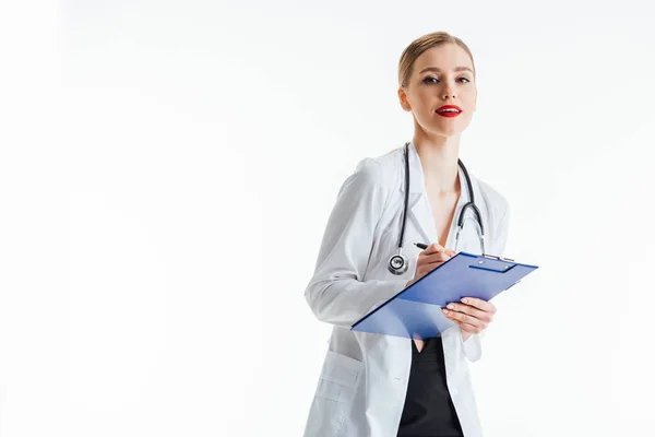 Glückliche und sexy Krankenschwester in weißem Mantel schreibt Rezept und hält Klemmbrett isoliert auf weiß — Stockfoto