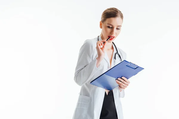 Infirmière coûteuse et sexy en manteau blanc regardant prescription tout en tenant presse-papiers et stylo près des lèvres rouges isolé sur blanc — Photo de stock