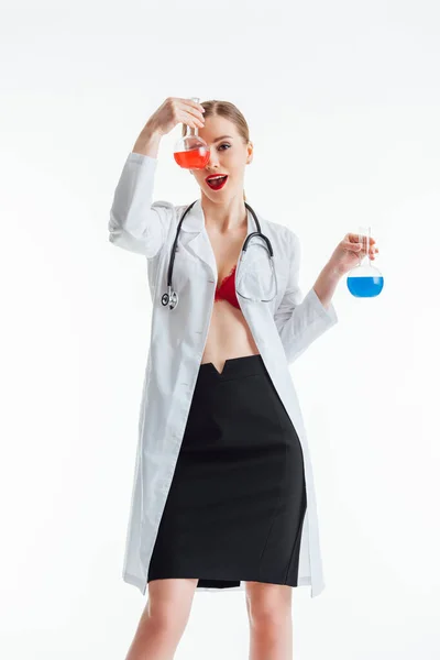 Infirmière gaie et sexy en manteau blanc tenant flacons avec liquide rouge et bleu isolé sur blanc — Photo de stock