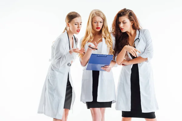 Sexy enfermeras en blanco abrigos gesto mientras mira portapapeles aislado en blanco - foto de stock