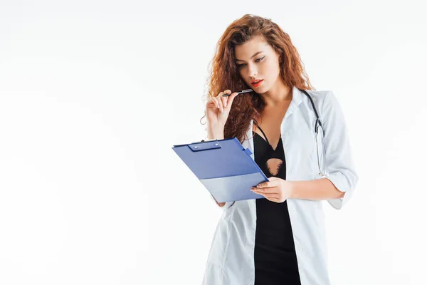 Infirmière bouclée et sexy tenant stylo et regardant presse-papiers isolé sur blanc — Photo de stock