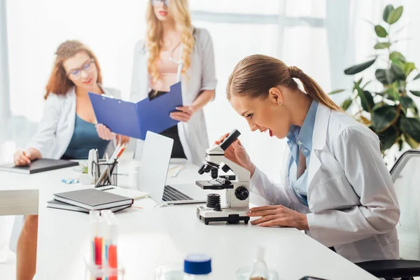 Selective focus of excited and sexy nurse looking through microscope near women — Stock Photo