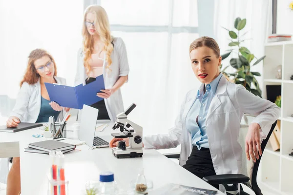 Selective focus of sexy nurse sitting near microscope and women — Stock Photo