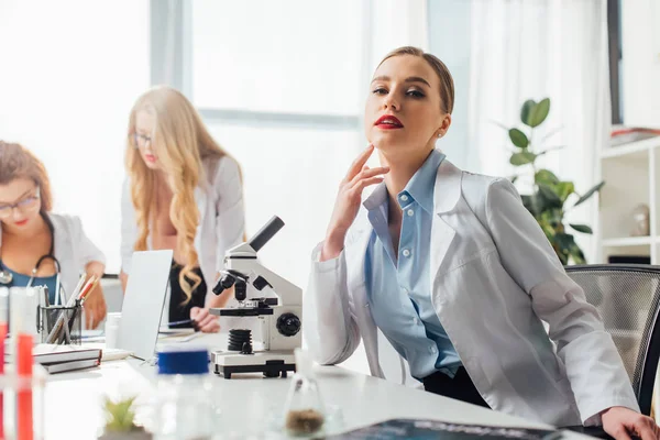 Selective focus of beautiful and sexy nurse sitting near microscope and women — Stock Photo