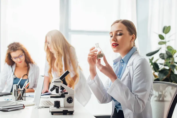 Selective focus of beautiful and sexy nurse looking at flask near women in white coats — Stock Photo