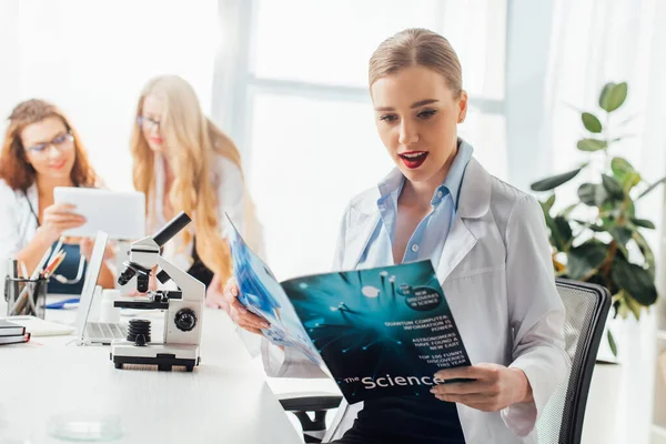 Selective focus of sexy nurse reading science magazine near women and microscope — Stock Photo