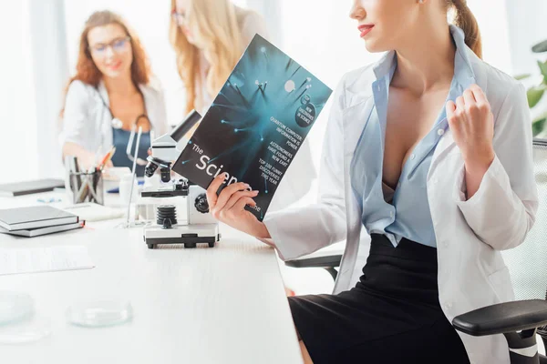 Enfoque selectivo de enfermera sexy celebración revista de ciencia cerca de las mujeres y microscopio - foto de stock