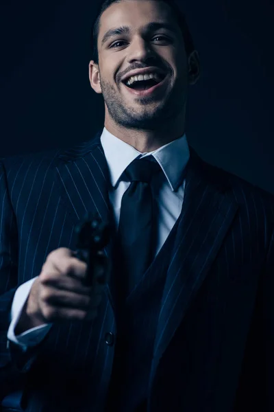 Selective focus of gangster aiming gun, laughing and looking at camera isolated on dark background — Stock Photo