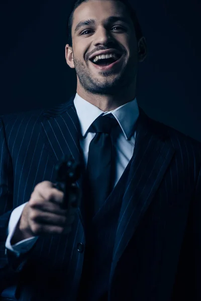 Focus selettivo del gangster mirare pistola, ridere e guardare la fotocamera isolata su sfondo scuro — Foto stock