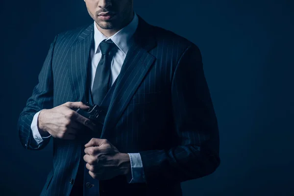 Cropped view of gangster hiding weapon in suit on dark blue background — Stock Photo