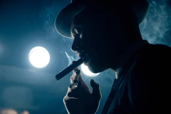 Vista de ángulo bajo del cigarro fumador mafioso sobre fondo azul oscuro - foto de stock