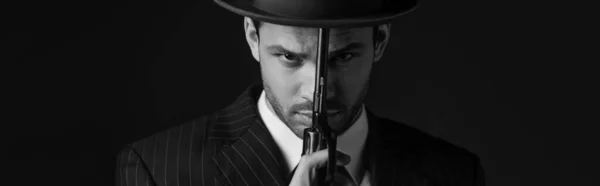 Imagen monocromática del gángster sosteniendo la pistola frente a la cara aislado en negro, tiro panorámico - foto de stock