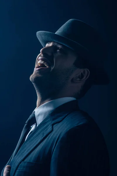 Mafioso en traje y sombrero de fieltro riendo sobre fondo azul oscuro - foto de stock