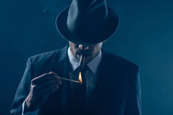 Mafioso avec yeux couverts avec chapeau feutre allumant cigare sur fond bleu foncé — Photo de stock