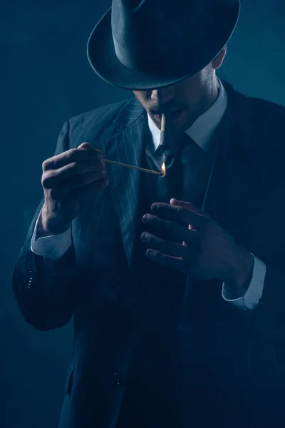 Mafioso con los ojos cubiertos con fieltro sombrero iluminación cigarro en azul oscuro - foto de stock