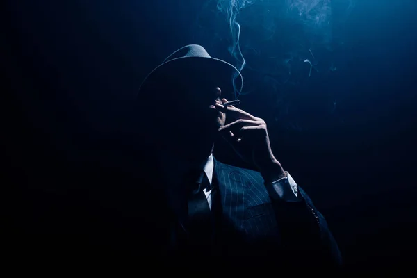 Silueta de mafioso en traje y fieltro sombrero fumar cigarrillo sobre fondo azul oscuro - foto de stock