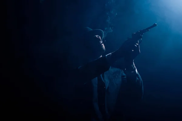 Silueta de gángster apuntando revólver y fumando sobre fondo oscuro - foto de stock