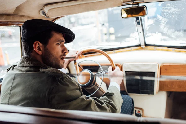 Selective focus of mafioso in coat and hat with cigarette holding steering wheel in car — Stock Photo