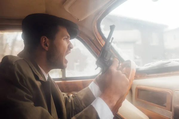 Profile of irritated gangster with gun shouting and driving car — Stock Photo