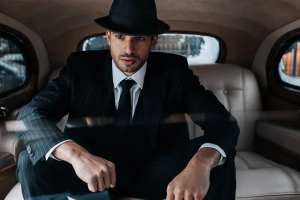 Selective focus of pensive mafioso in black suit and felt hat in retro car — Stock Photo