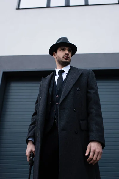 Low angle view of gangster in black coat and hat with weapon on street — Stock Photo