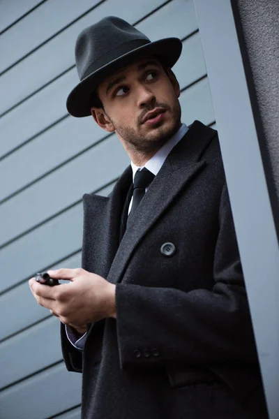 Low angle view of mafioso holding gun and looking away in ambush near wall on street — Stock Photo