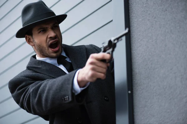 Selective focus of mafioso aiming gun with outstretched hand and shouting from corner near wall on street — Stock Photo