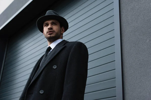 Low angle view of mafioso in black coat and felt hat near wall on street — Stock Photo