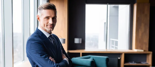 Panoramic shot of smiling businessman in suit with crossed arms looking at camera in hotel — Stock Photo