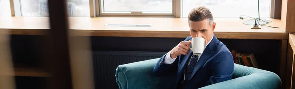 Panoramic shot of businessman in suit drinking coffee and sitting in armchair — Stock Photo