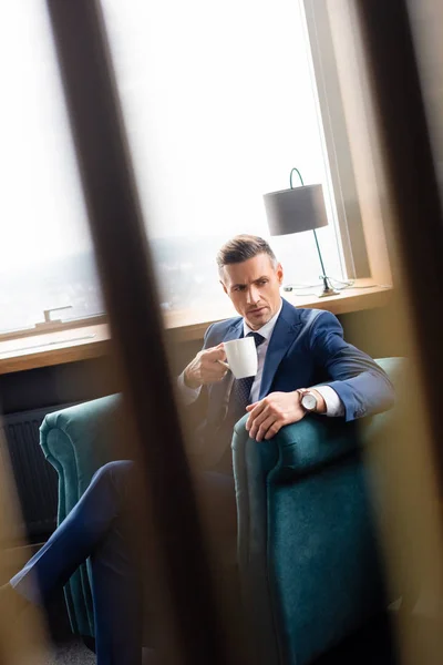 Enfoque selectivo de hombre de negocios en traje sosteniendo taza y sentado en sillón - foto de stock