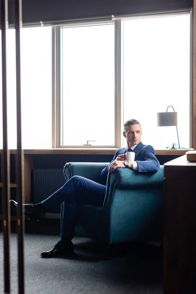 Geschäftsmann im Anzug sitzt im Sessel mit Tasse und schaut weg — Stockfoto