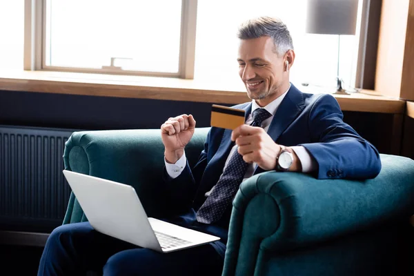 Homme d'affaires souriant en costume tenant la carte de crédit et en utilisant un ordinateur portable — Photo de stock