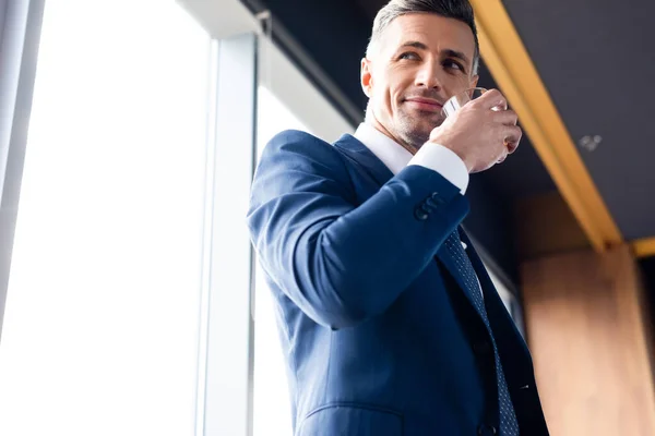 Vista de ángulo bajo del hombre de negocios sonriente en traje sosteniendo vidrio - foto de stock