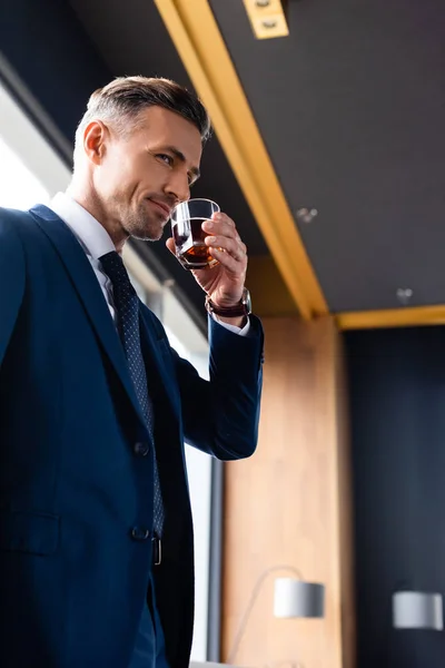 Bajo ángulo vista de sonriente hombre de negocios en traje de beber coñac - foto de stock