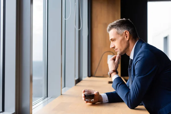 Side view of pensive businessman in suit holding glass and looking through window — Stock Photo