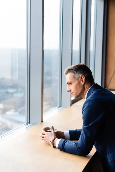 Vista lateral del hombre de negocios en traje sosteniendo vidrio y mirando a través de la ventana — Stock Photo