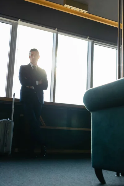 Low angle view of businessman in suit with crossed arms standing near travel bag — Stock Photo