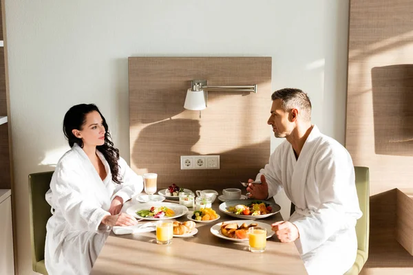 Vista lateral do namorado e namorada em roupões de banho conversando e tomando café da manhã — Fotografia de Stock