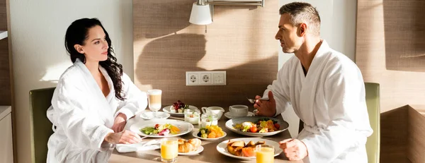 Panoramic shot of boyfriend and girlfriend in bathrobes talking and having breakfast — Stock Photo