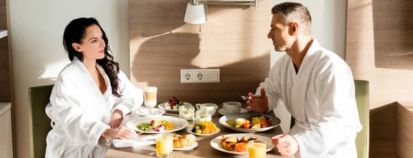 Plan panoramique de petit ami et petite amie en peignoirs parler et prendre le petit déjeuner — Photo de stock