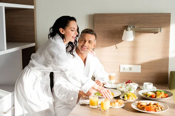 Petit ami souriant et petite amie en peignoirs prendre le petit déjeuner à l'hôtel — Photo de stock