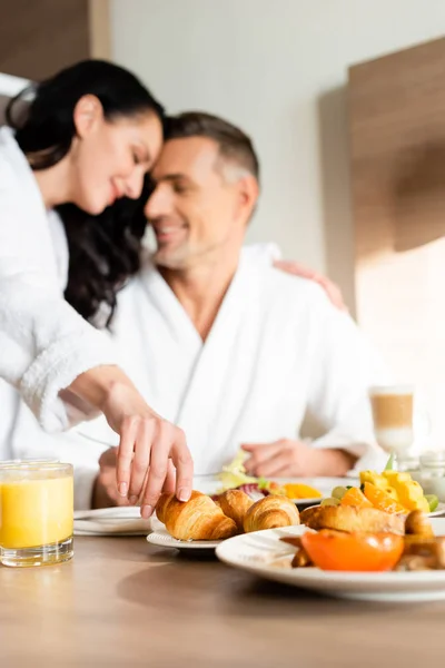 Selective focus of smiling girlfriend in bathrobe hugging boyfriend and taking croissant in hotel — Stock Photo