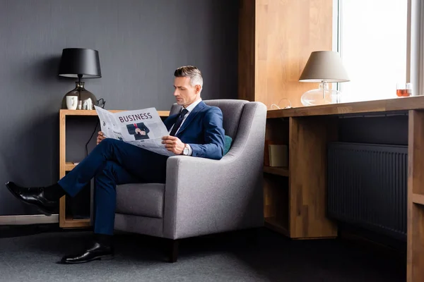 Hombre de negocios en traje leyendo periódico con letras de negocios y sentado en sillón - foto de stock