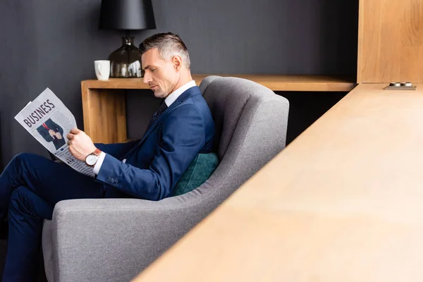 Вид сбоку бизнесмена в костюме, читающего газету с деловым уклоном и сидящего в кресле — стоковое фото