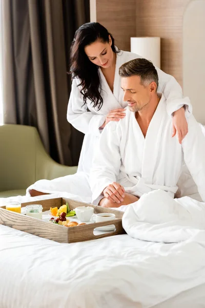 Smiling girlfriend hugging boyfriend and looking at food on tray in hotel — Stock Photo