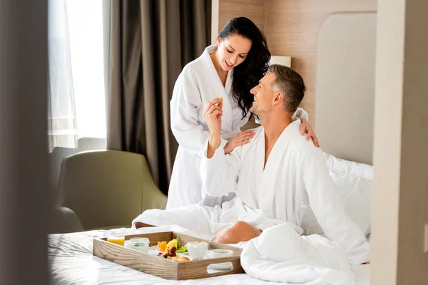 Sonriente novio alimentación atractiva novia con uva en hotel - foto de stock