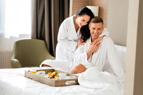 Novia sonriente en albornoz abrazando novio guapo en el hotel - foto de stock