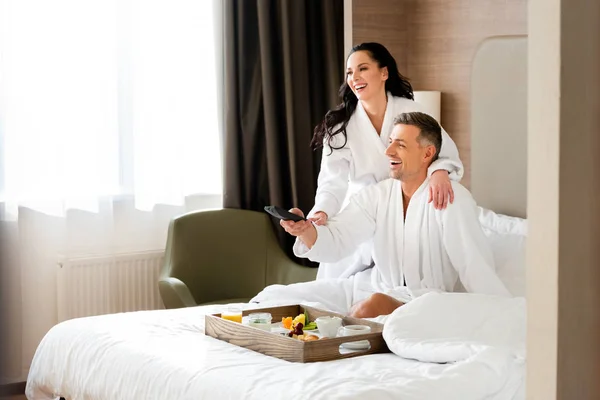 Lächelnde Freundin im Bademantel umarmt schönen Freund mit Fernbedienung im Hotel — Stockfoto